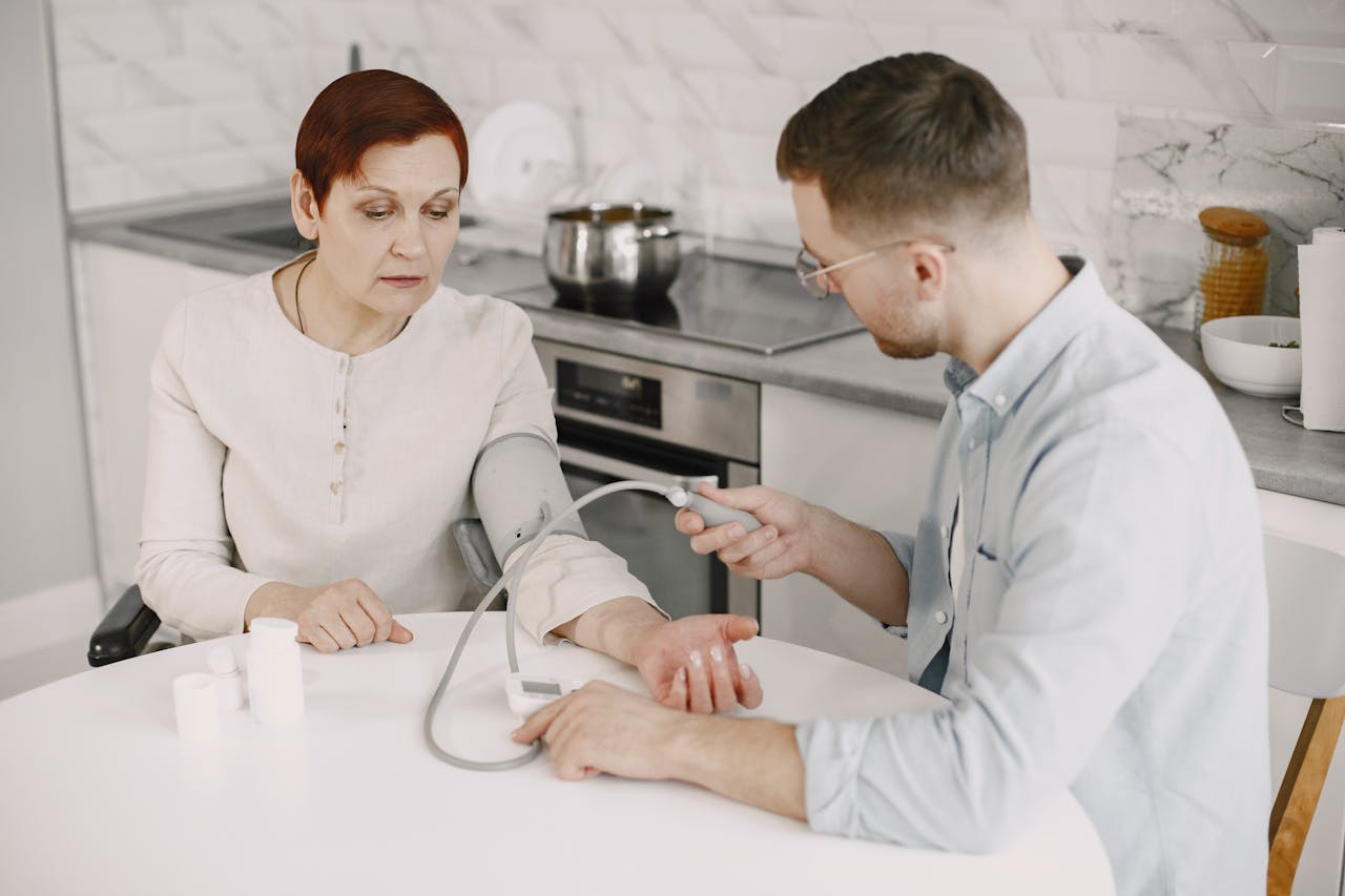 Adult man monitoring elderly womans blood pressure at kitchen table using sphygmomanometer.