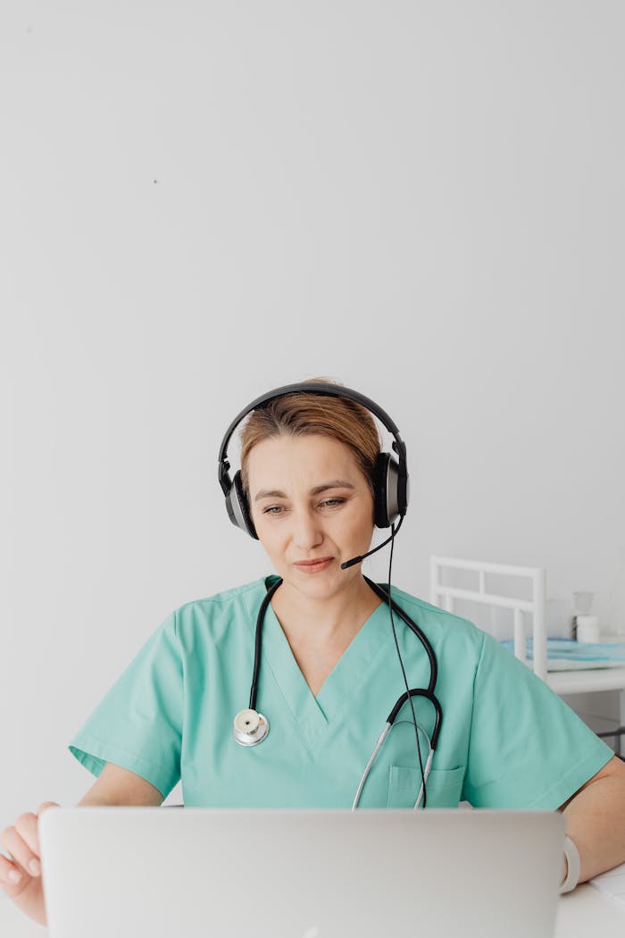 A healthcare worker in medical scrubs using a laptop and headset for telemedicine.